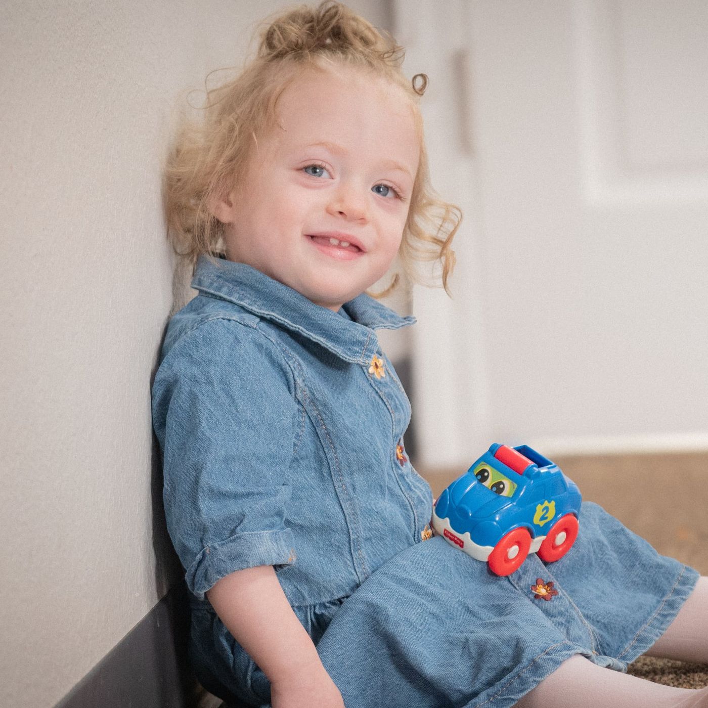 A smiling little girl in a denim dress holding a toy car
